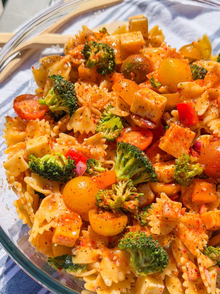 A close-up of The Best Pasta Salad featuring bowtie pasta, broccoli florets, cherry tomatoes, cheese cubes, and red bell peppers, all seasoned with herbs and spices in a glass bowl.