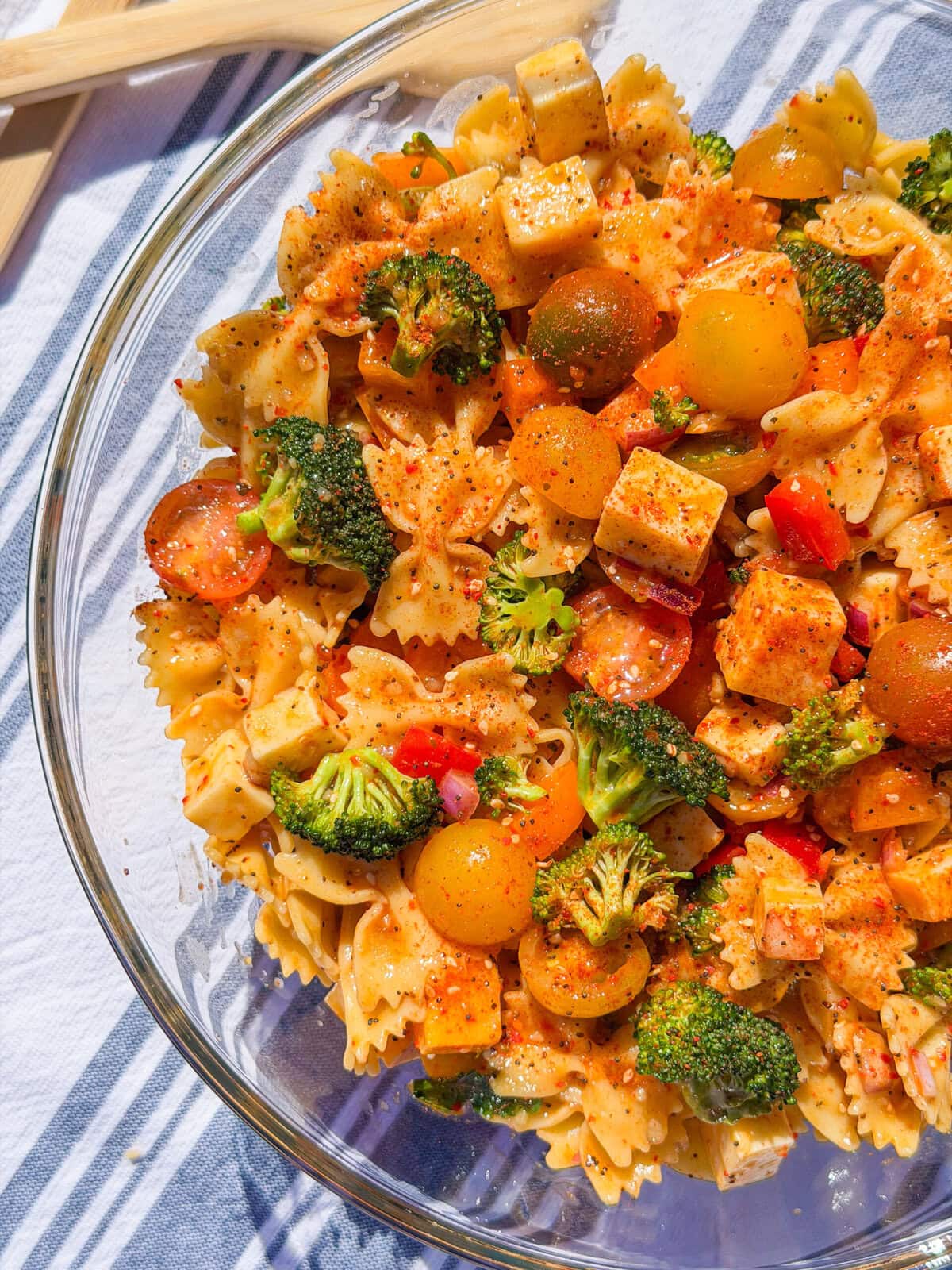 A glass bowl filled with The Best Pasta Salad, featuring bowtie pasta, broccoli, cherry tomatoes, red bell pepper, and cubes of cheese tossed with spices. Wooden utensils rest in the background on a striped cloth.