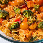 A close-up of The Best Pasta Salad featuring bowtie pasta, broccoli, yellow cherry tomatoes, cubed cheese, red onions, and a sprinkle of seasoning, all tossed in a light dressing.