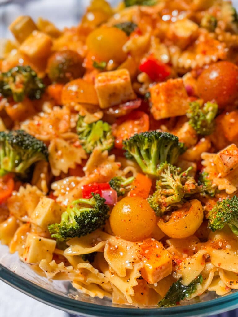 A close-up of The Best Pasta Salad featuring bowtie pasta, broccoli, yellow cherry tomatoes, cubed cheese, red onions, and a sprinkle of seasoning, all tossed in a light dressing.