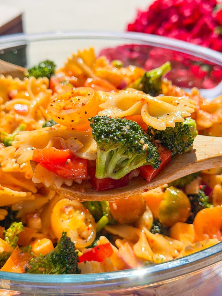 A close-up of a wooden spoon holding The Best Pasta Salad with bowtie pasta, broccoli, cherry tomatoes, and red bell peppers in a glass bowl, set against a bright, colorful background.