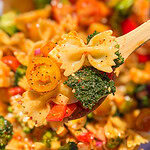 A wooden spoon holds bowtie pasta salad—The Best Pasta Salad—brimming with broccoli, cherry tomatoes, red bell peppers, and a light dressing, with more vibrant salad in the background.