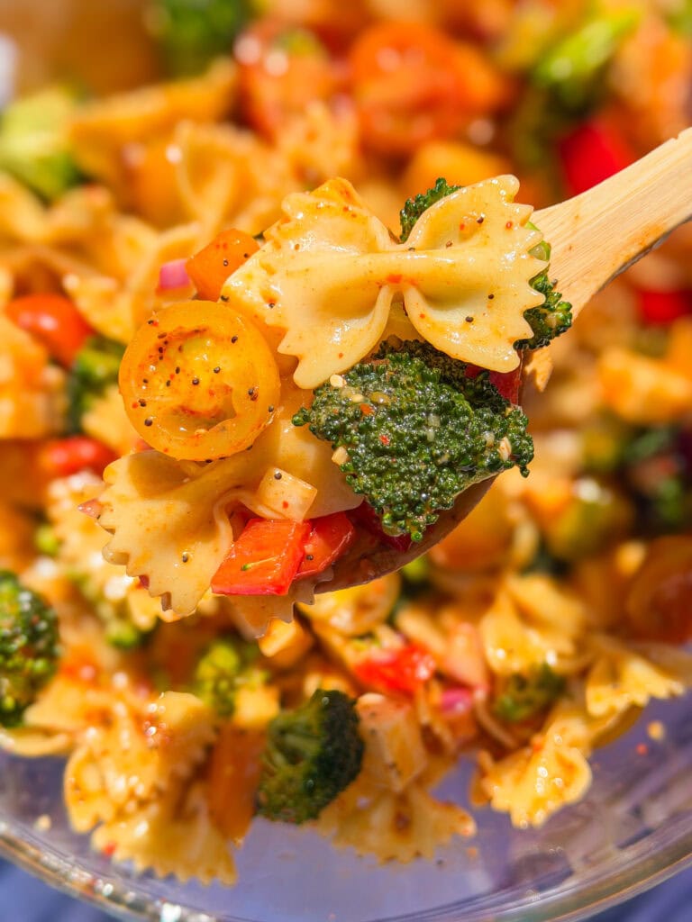 A wooden spoon holds bowtie pasta salad—The Best Pasta Salad—brimming with broccoli, cherry tomatoes, red bell peppers, and a light dressing, with more vibrant salad in the background.