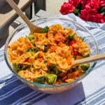 A glass bowl holds The Best Pasta Salad, featuring bowtie pasta, broccoli, cherry tomatoes, cheese cubes, and red pepper tossed in dressing. Wooden spoons rest in the bowl on a striped cloth beside bright pink flowers.