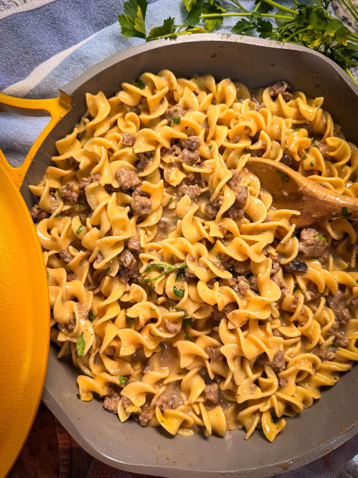 A skillet filled with creamy egg noodles and ground beef, resembling a Homemade Hamburger Helper Beef Stroganoff, is garnished with parsley. A wooden spoon rests in the pan, with fresh parsley visible in the background.