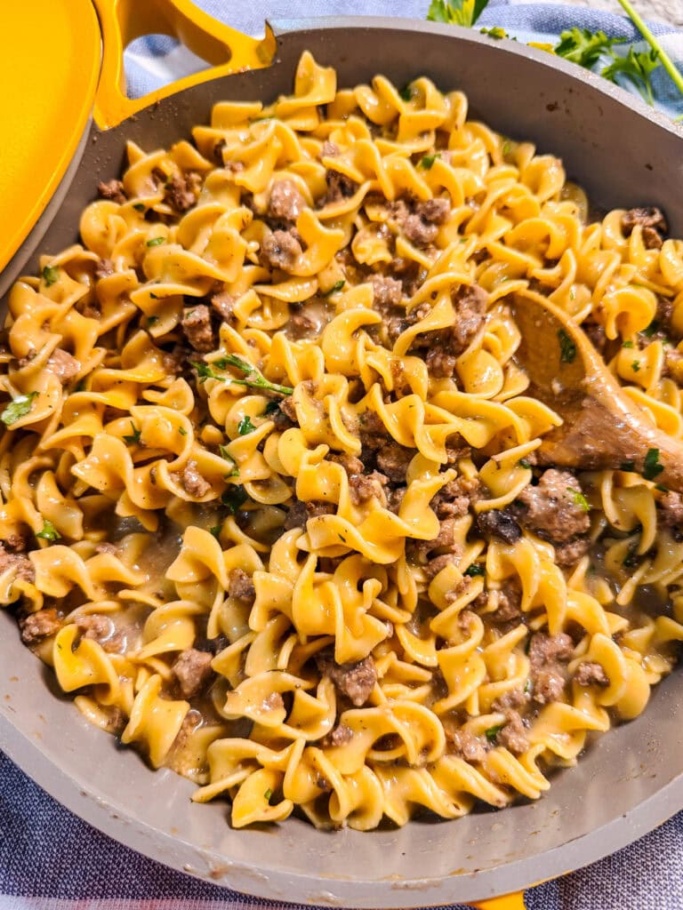 A skillet filled with cooked egg noodles and ground beef, creating a homemade Hamburger Helper Beef Stroganoff, is garnished with chopped herbs. A wooden spoon stirs the mixture, while a yellow lid peeks in from the top left corner.