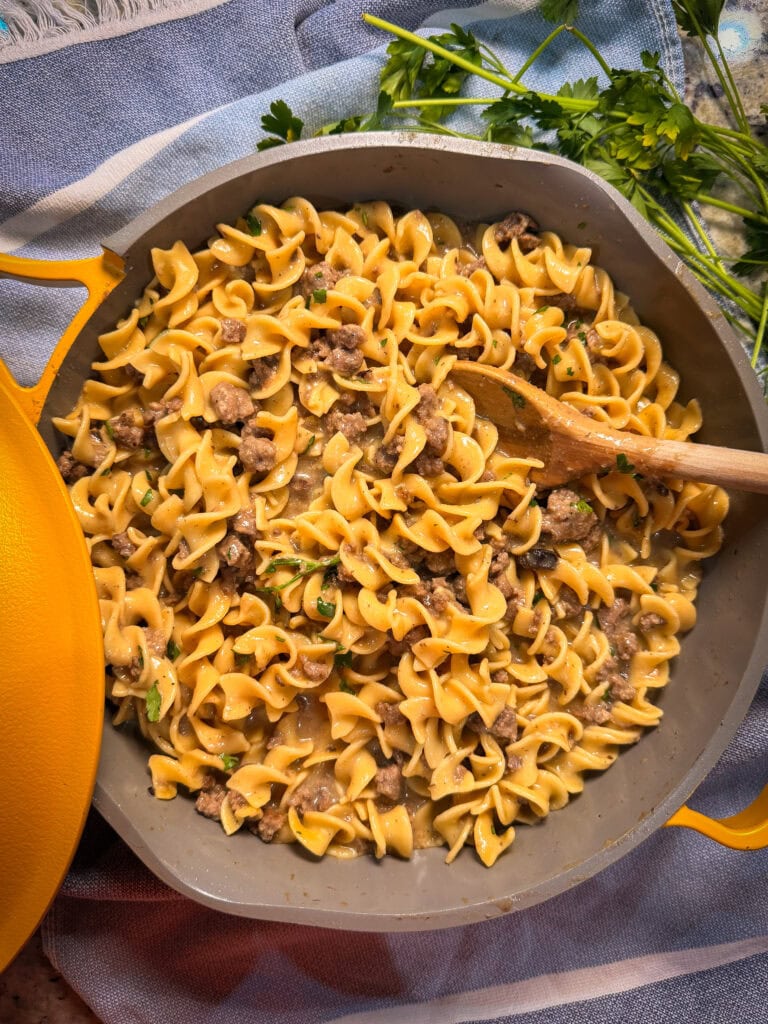 A skillet filled with egg noodles, ground beef, and a creamy sauce, garnished with chopped parsley—this Homemade Hamburger Helper Beef Stroganoff looks inviting. A wooden spoon rests in the pan, with fresh parsley on a blue and white cloth nearby.
