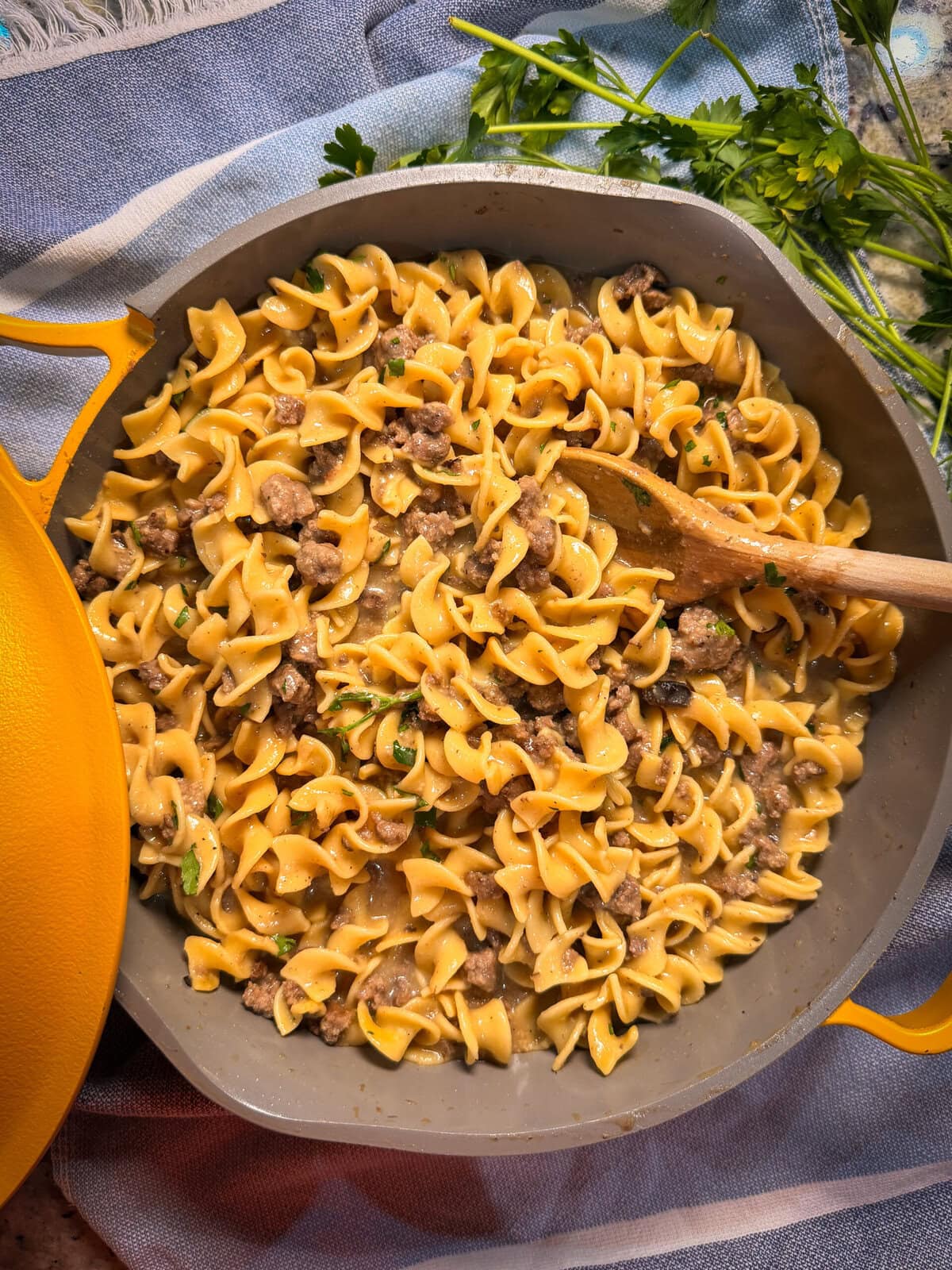 A skillet filled with egg noodles, ground beef, and a creamy sauce, garnished with chopped parsley—this Homemade Hamburger Helper Beef Stroganoff looks inviting. A wooden spoon rests in the pan, with fresh parsley on a blue and white cloth nearby.