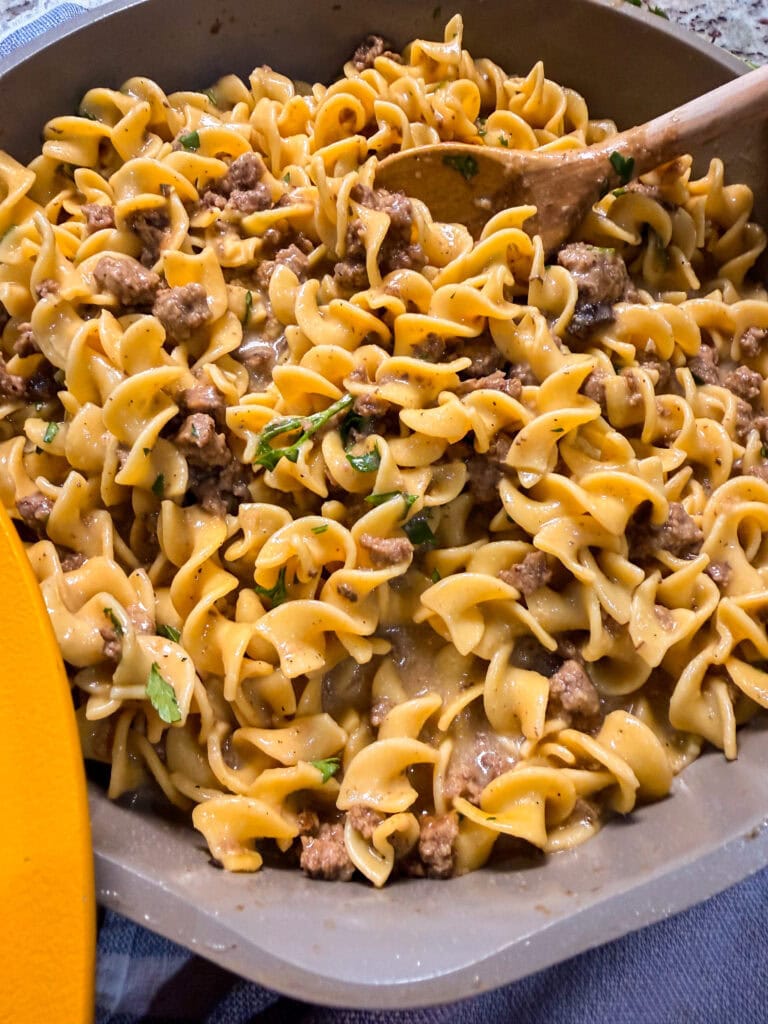 A close-up of a skillet filled with creamy Homemade Hamburger Helper Beef Stroganoff, garnished with chopped herbs. A wooden spoon rests in the dish, where spiral noodles and ground beef mingle in a rich sauce.