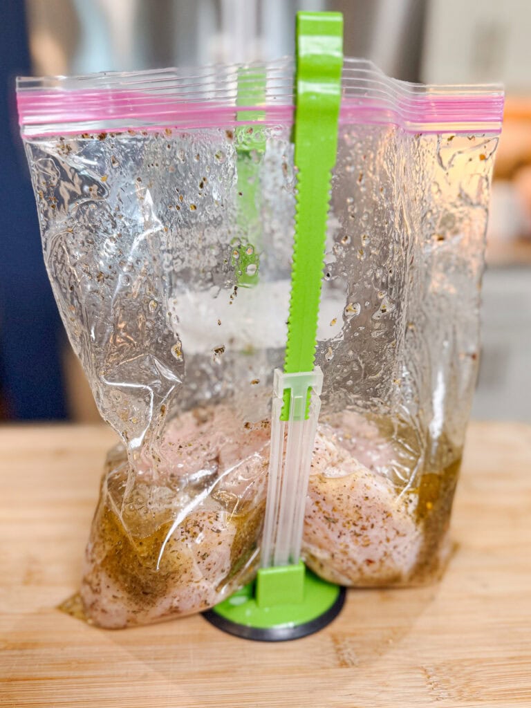 Raw chicken pieces for Sheet Pan Italian Style Chicken marinating in a sealed plastic bag, which is held upright by a green bag holder on a wooden surface in a kitchen setting.