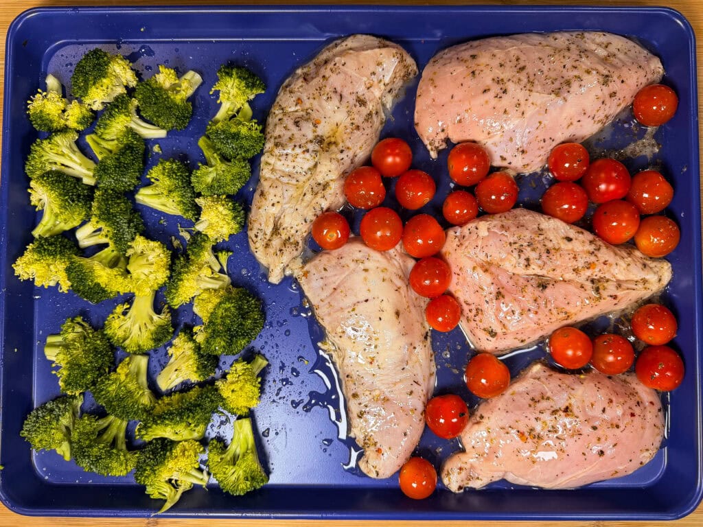 A blue baking tray with raw Sheet Pan Italian Style Chicken breasts, cherry tomatoes, and broccoli florets arranged separately, ready for roasting.