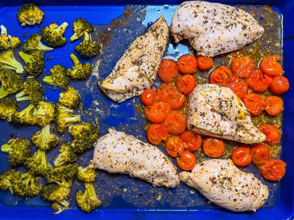 Sheet Pan Italian Style Chicken breasts seasoned with herbs, surrounded by roasted cherry tomatoes and broccoli florets on a blue baking tray.