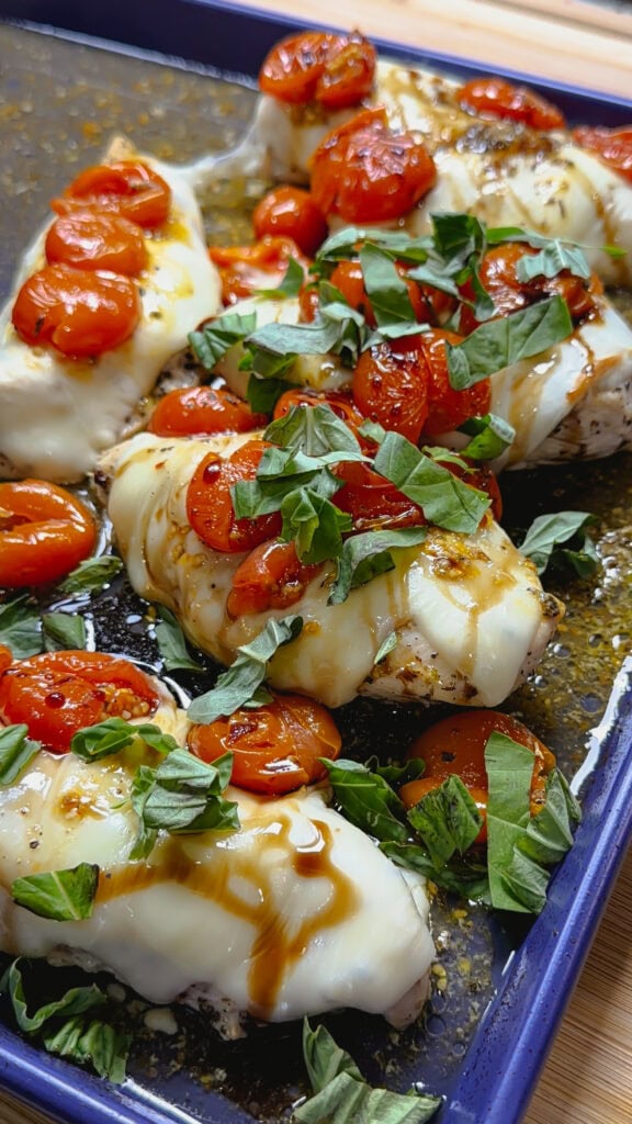 Sheet Pan Italian Style Chicken features baked chicken breasts topped with melted cheese, roasted cherry tomatoes, fresh basil, and a drizzle of balsamic glaze on a blue baking tray.