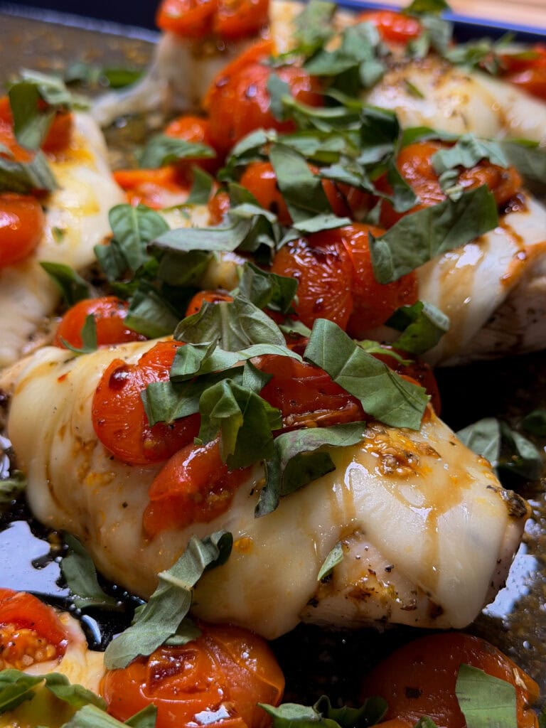 Sheet Pan Italian Style Chicken breasts are baked to perfection, then topped with melted cheese, roasted cherry tomatoes, and fresh chopped basil leaves—all prepared conveniently on a baking sheet.