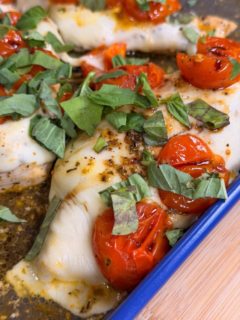 Sheet Pan Italian Style Chicken: Baked chicken breasts topped with roasted cherry tomatoes, chopped fresh basil, and seasonings, shown in a blue baking dish on a wooden surface.