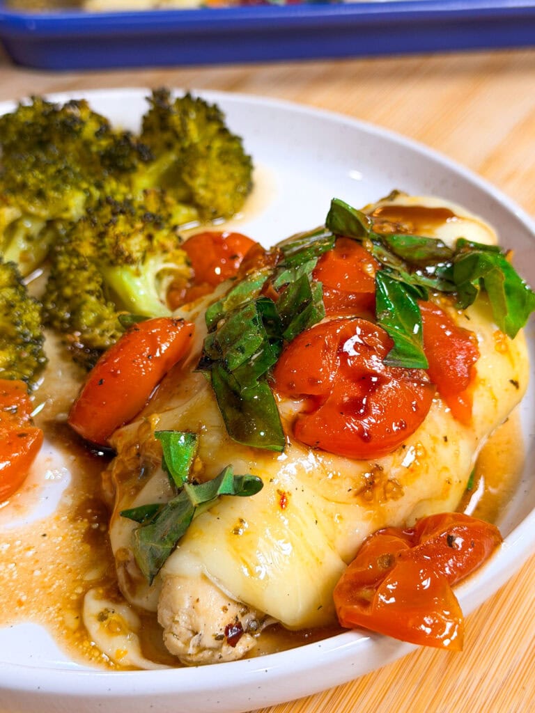 A plate of Sheet Pan Italian Style Chicken breast topped with melted cheese, cherry tomatoes, and fresh basil, served with roasted broccoli on the side.