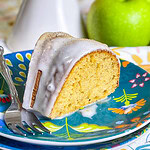 Golden brown apple cider Bundt cake drizzled with cider glaze and sprinkled with cinnamon sugar on a white cake stand.