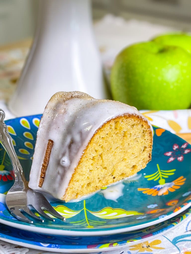 Golden brown apple cider Bundt cake drizzled with cider glaze and sprinkled with cinnamon sugar on a white cake stand.