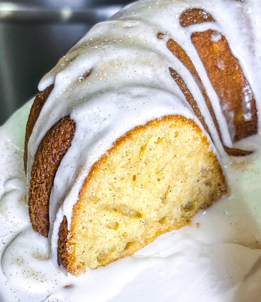 Golden brown apple cider Bundt cake drizzled with cider glaze and sprinkled with cinnamon sugar on a white cake stand.