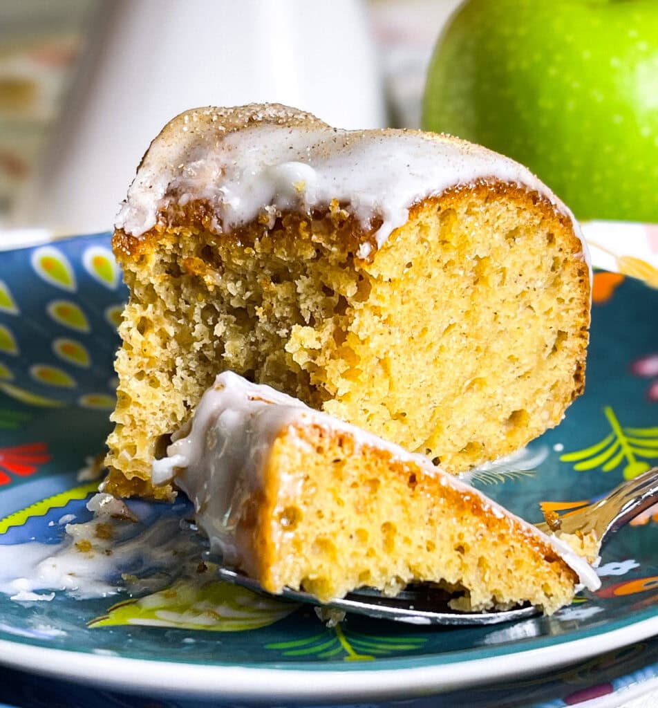 Golden brown apple cider Bundt cake drizzled with cider glaze and sprinkled with cinnamon sugar on a white cake stand.