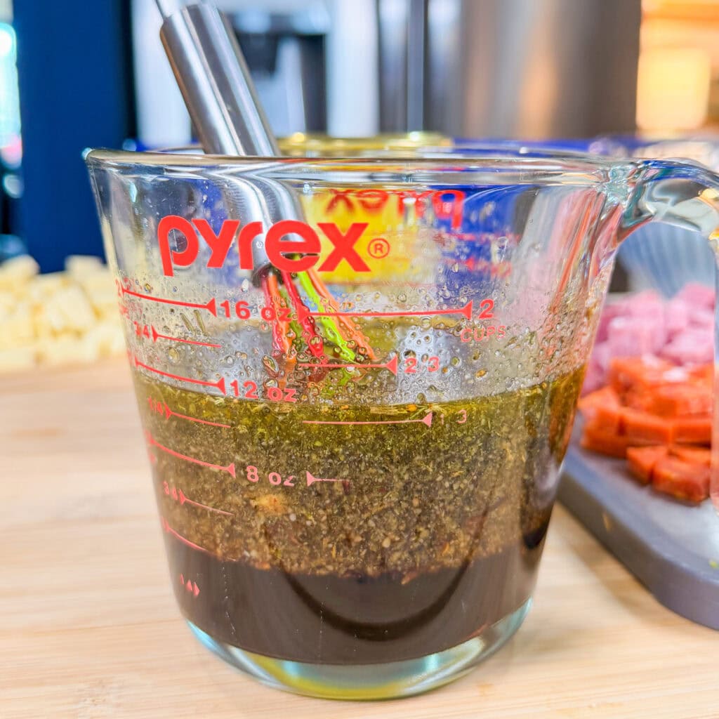 A Pyrex measuring cup filled with a dark brown liquid and seasonings, being stirred with a metal whisk. In the background, diced ingredients for an Italian Sub Pasta Salad are visible on a cutting board.