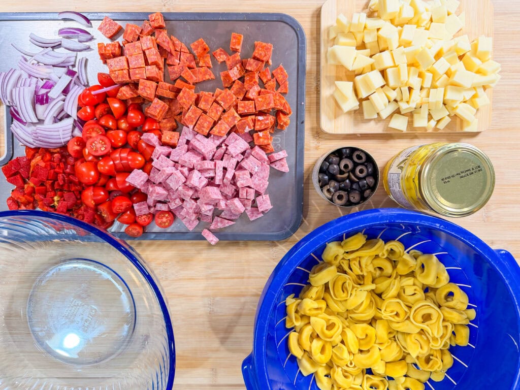 A wooden surface with bowls and trays holding ingredients for an Italian Sub Pasta Salad: sliced red onions, cherry tomatoes, diced meats, cubed cheese, black olives, a jar of yellow peppers, and cooked tortellini in a blue strainer.