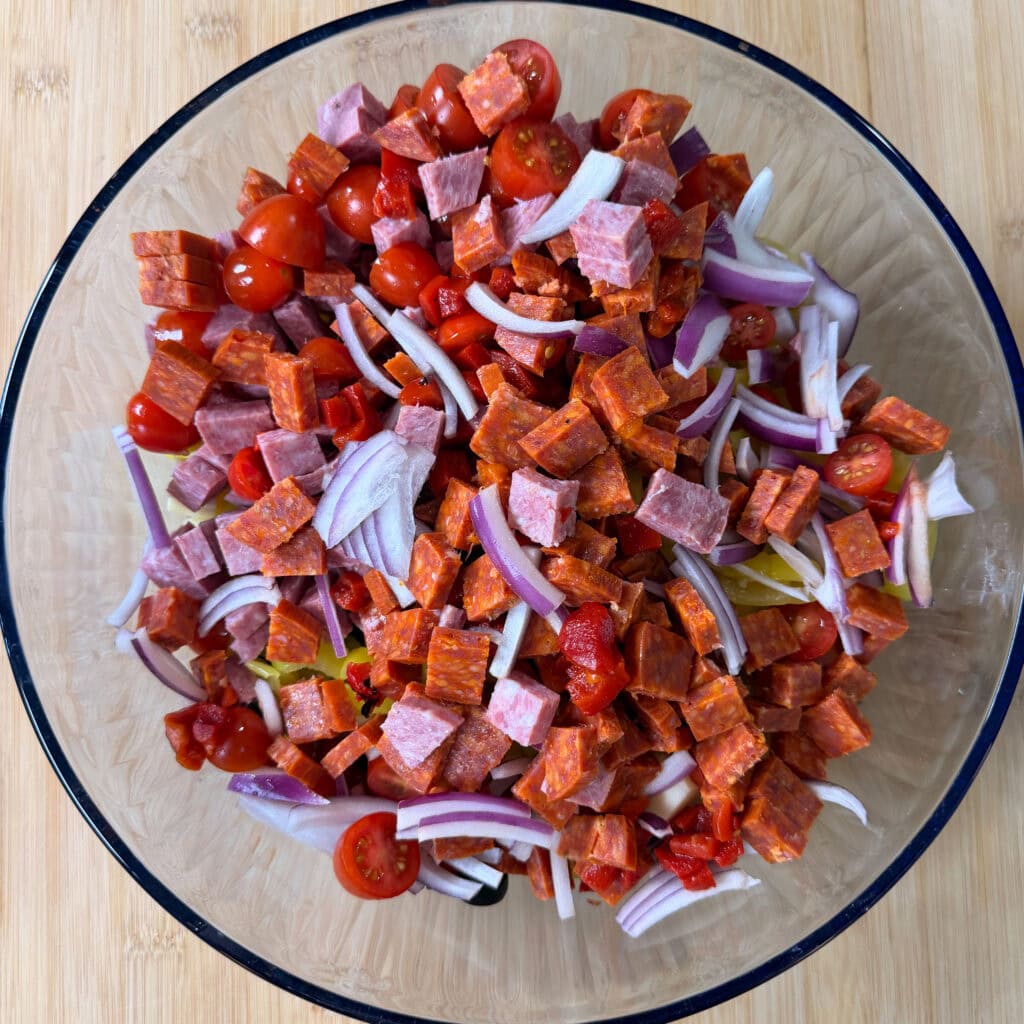A glass bowl filled with Italian Sub Pasta Salad—sliced cherry tomatoes, chopped pepperoni, cubed salami, thinly sliced red onions, and chopped peppers—placed on a wooden surface.