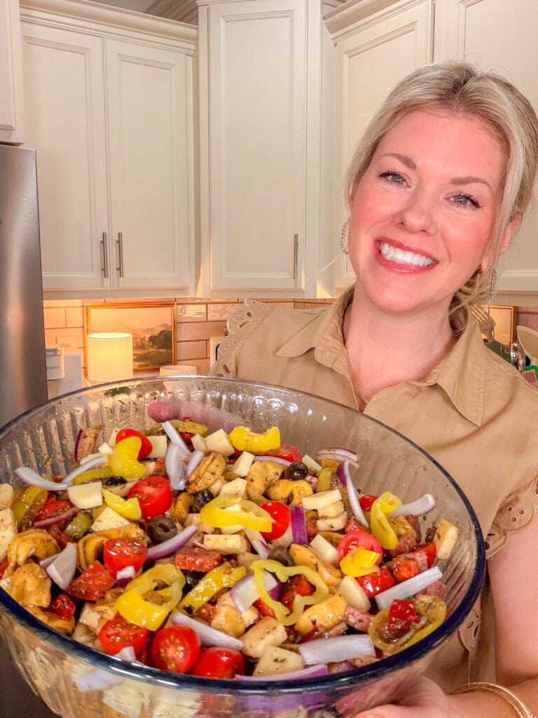 A smiling woman in a tan shirt holds a large glass bowl filled with colorful Italian Sub Pasta Salad, featuring cherry tomatoes, yellow peppers, onions, and pasta pieces, in a bright kitchen.