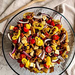 A glass bowl filled with Italian Sub Pasta Salad featuring tortellini, cherry tomatoes, red onion slices, cheese cubes, olives, and pepperoncini peppers, with a wooden spoon and towel in the background.