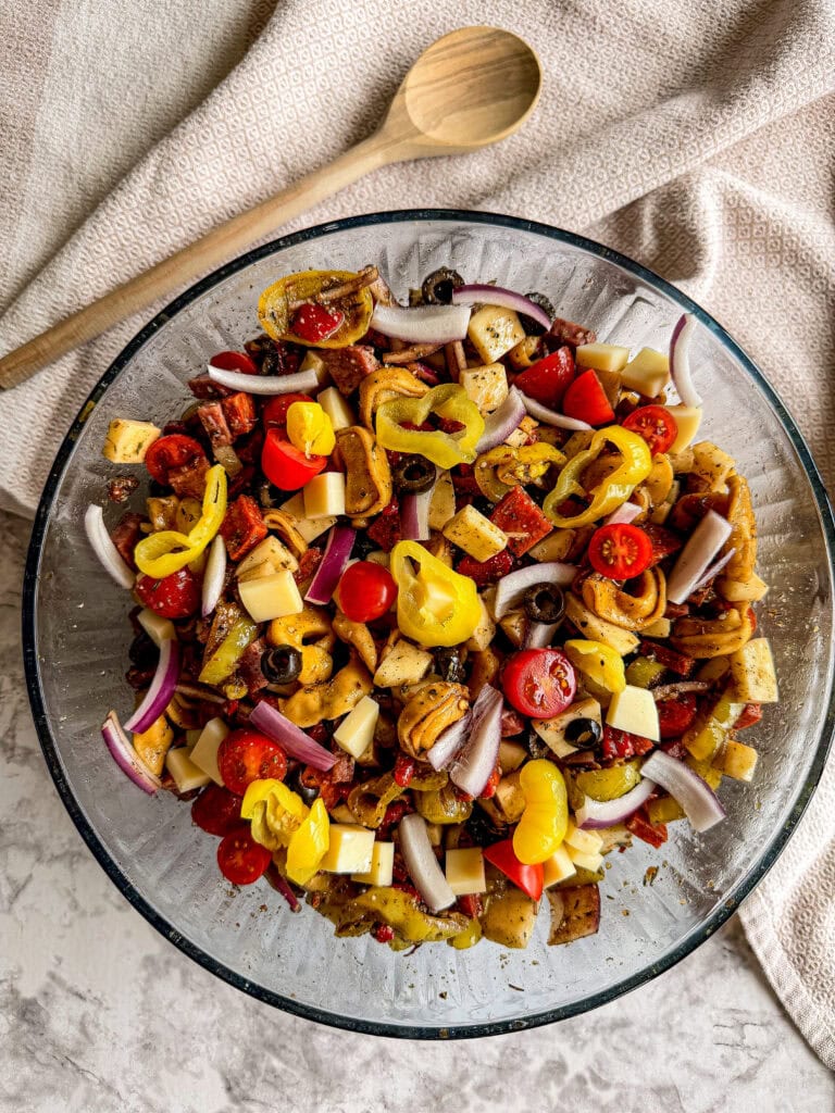A glass bowl filled with Italian Sub Pasta Salad featuring tortellini, cherry tomatoes, red onion slices, cheese cubes, olives, and pepperoncini peppers, with a wooden spoon and towel in the background.