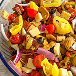 A close-up of an Italian Sub Pasta Salad in a bowl with rotini pasta, cherry tomatoes, cubed cheese, black olives, red onion, pepperoni, banana peppers, and Italian seasoning.