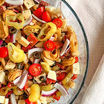A glass bowl filled with Italian Sub Pasta Salad, featuring tortellini, cherry tomatoes, red onion slices, cheese cubes, olives, pepperoncini, and Italian seasoning. A beige textured towel sits beside the bowl on a light surface.