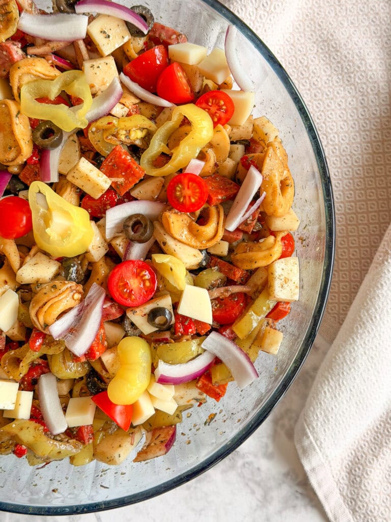 A glass bowl filled with Italian Sub Pasta Salad, featuring tortellini, cherry tomatoes, red onion slices, cheese cubes, olives, pepperoncini, and Italian seasoning. A beige textured towel sits beside the bowl on a light surface.