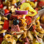 A wooden spoon holds a serving of Italian Sub Pasta Salad with tortellini, salami, olives, red onions, cherry tomatoes, peppers, and cheese, with more salad blurred in the background.
