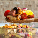 A wooden spoon holds a colorful mix of Italian Sub Pasta Salad with chunks of meat, cherry tomatoes, olives, red onion, and yellow bell peppers above a glass bowl filled with more salad.