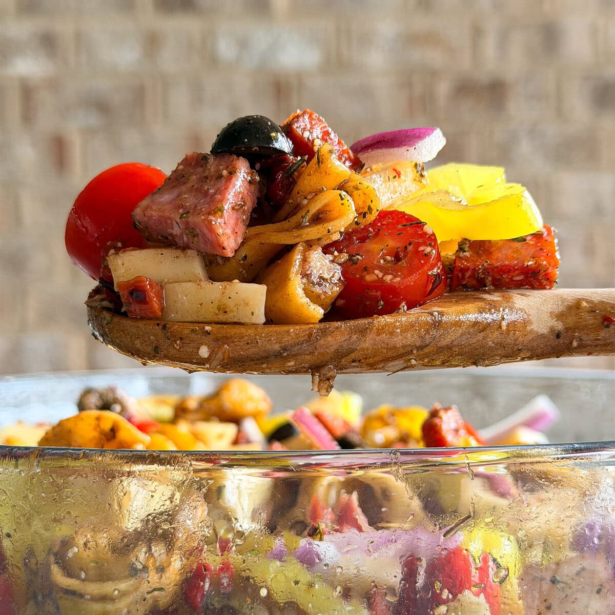 A wooden spoon holds a colorful mix of Italian Sub Pasta Salad with chunks of meat, cherry tomatoes, olives, red onion, and yellow bell peppers above a glass bowl filled with more salad.