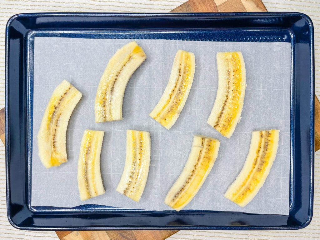 Sliced plantains arranged on a parchment-lined baking sheet, ready to be baked like homemade Frozen Banana Treats. The baking sheet sits on a wooden surface.