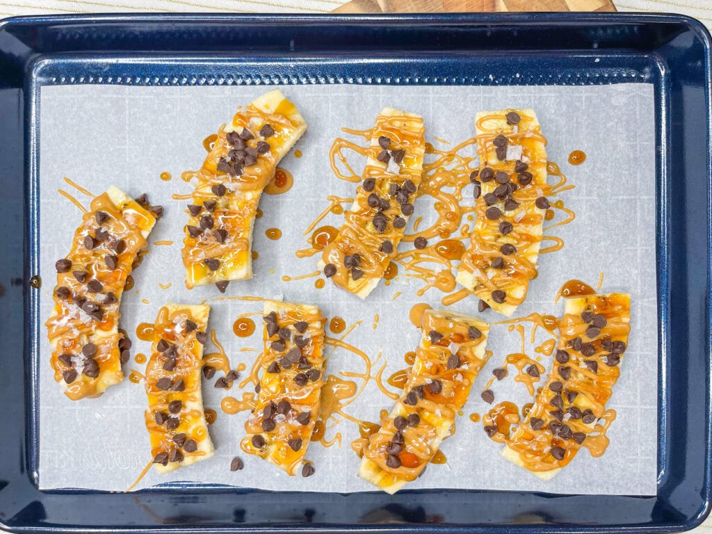 Frozen Banana Treats: Sliced bananas on a baking tray lined with parchment paper, topped with caramel sauce and chocolate chips, arranged in rows and ready to be baked or served.