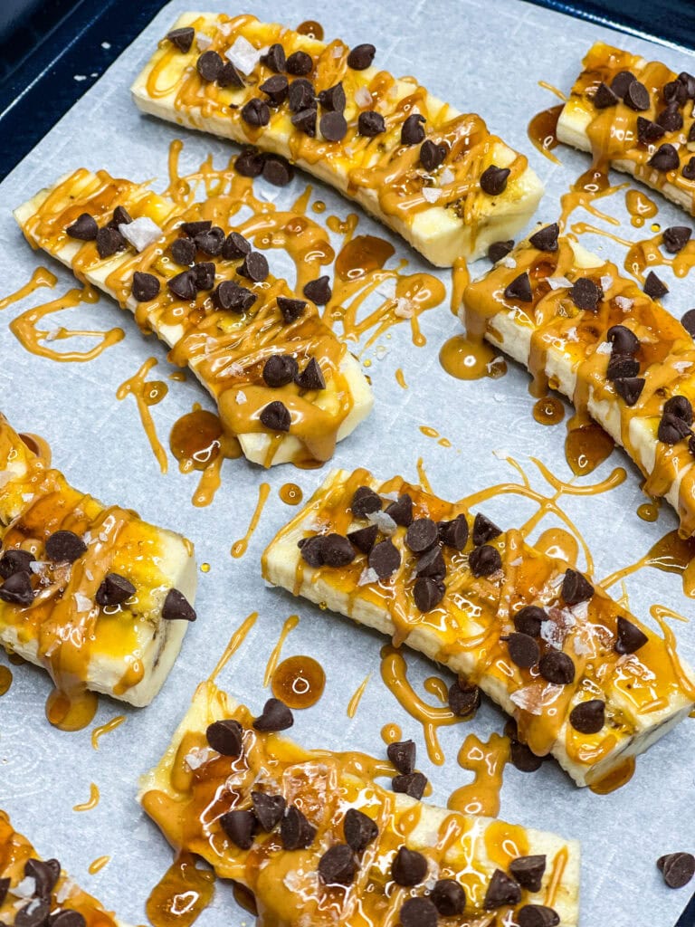 Sliced bananas on parchment paper topped with caramel sauce and scattered chocolate chips, these Frozen Banana Treats are ready to be baked or served as a sweet treat.
