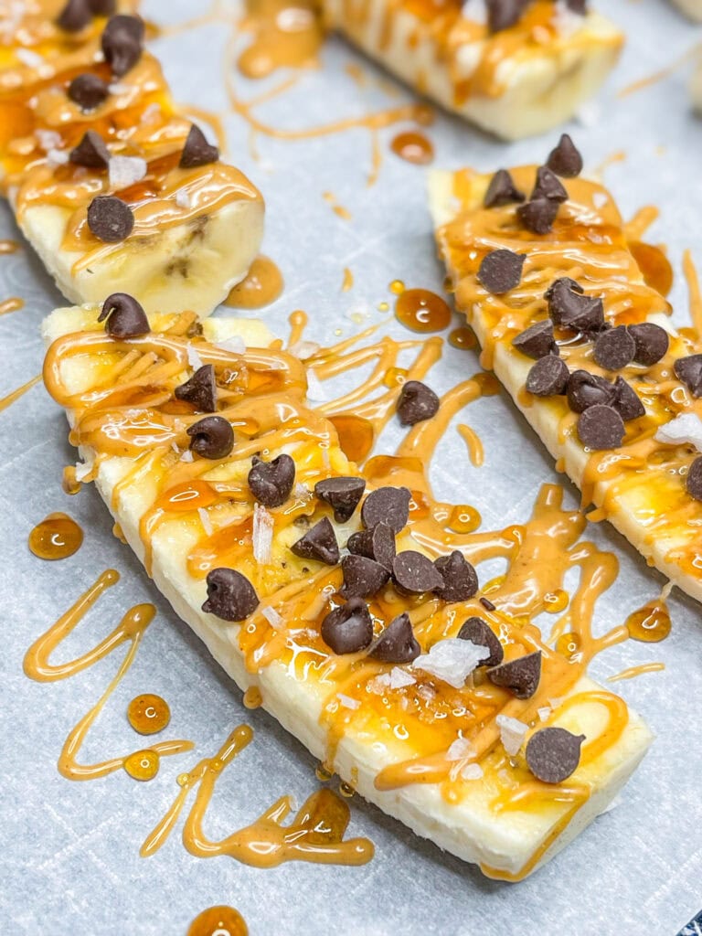Frozen Banana Treats: Sliced bananas topped with peanut butter, chocolate chips, and a drizzle of caramel sauce are arranged on parchment paper.