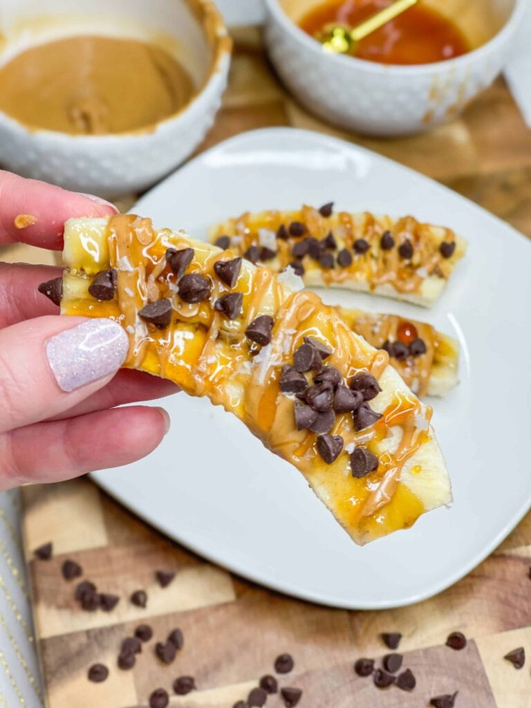 A hand holds a Frozen Banana Treat topped with peanut butter, chocolate chips, and honey. Two more of these tasty banana slices sit on a white plate; bowls of peanut butter and honey are in the background on a wooden surface.