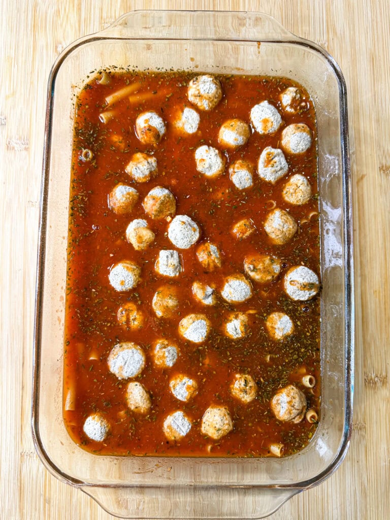 A glass baking dish filled with tomato-based sauce, meatballs, herbs, and a few pieces of pasta sits on a light wooden surface—perfect for serving as a hearty One Dish Meatball Casserole.