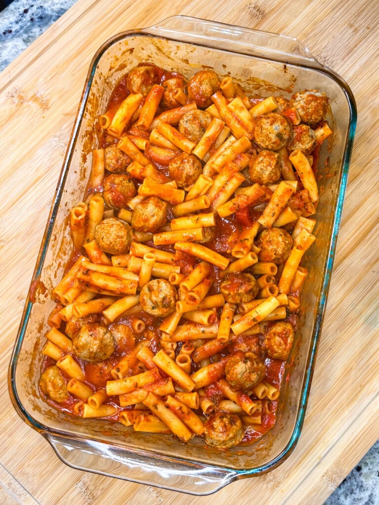 A glass baking dish filled with One Dish Meatball Casserole sits on a wooden cutting board, featuring baked ziti pasta, meatballs, and tomato sauce mixed together and perfectly coated for a hearty, comforting meal.