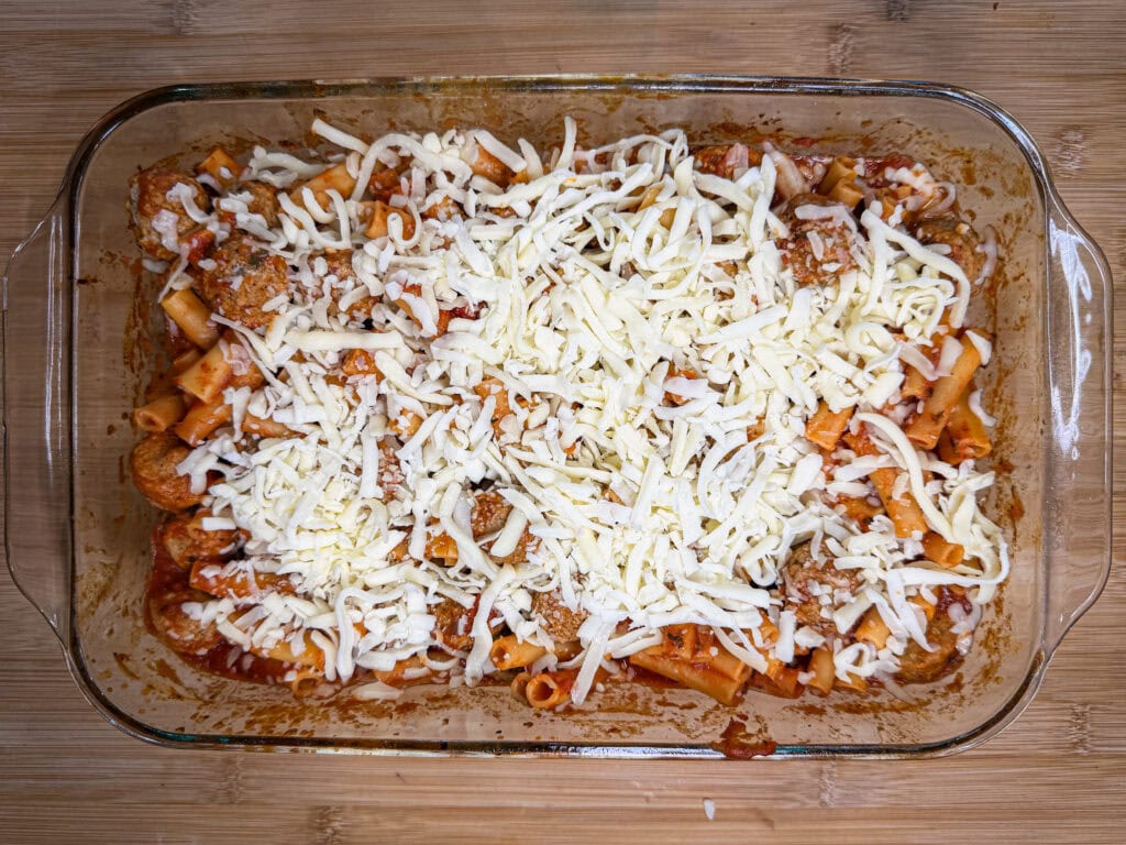A glass baking dish filled with One Dish Meatball Casserole—ziti pasta, tomato sauce, hearty meatballs, and a generous layer of shredded mozzarella—sits on a wooden surface.