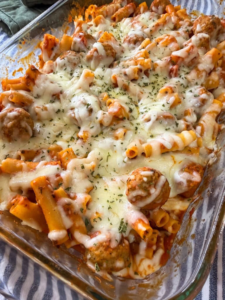 A glass baking dish filled with One Dish Meatball Casserole—ziti pasta, meatballs, marinara sauce, and melted cheese topped with herbs. The cheesy casserole is hot and fresh, sitting on a blue-and-white striped cloth.