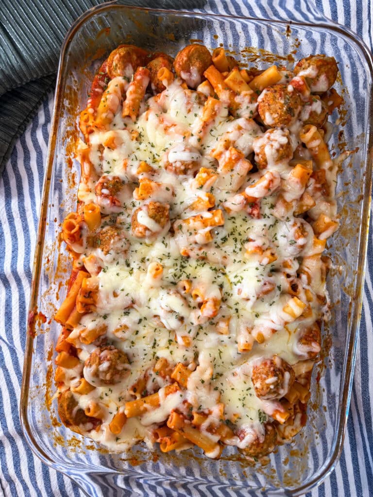 A glass baking dish filled with One Dish Meatball Casserole—baked ziti mixed with meatballs, tomato sauce, and topped with melted cheese and herbs—rests on a blue and white striped cloth.