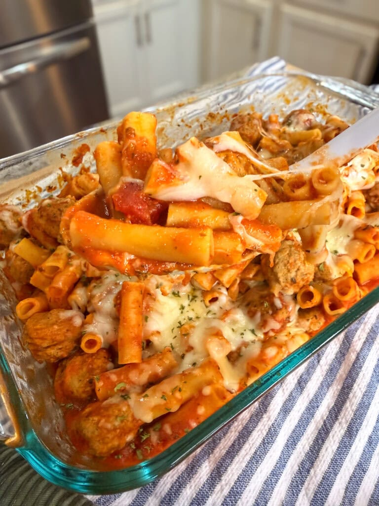 A glass baking dish filled with One Dish Meatball Casserole—baked ziti, meatballs, tomato sauce, and melted cheese—sits on a striped towel as a serving spoon lifts a portion. Kitchen cabinets and an oven are visible in the background.
