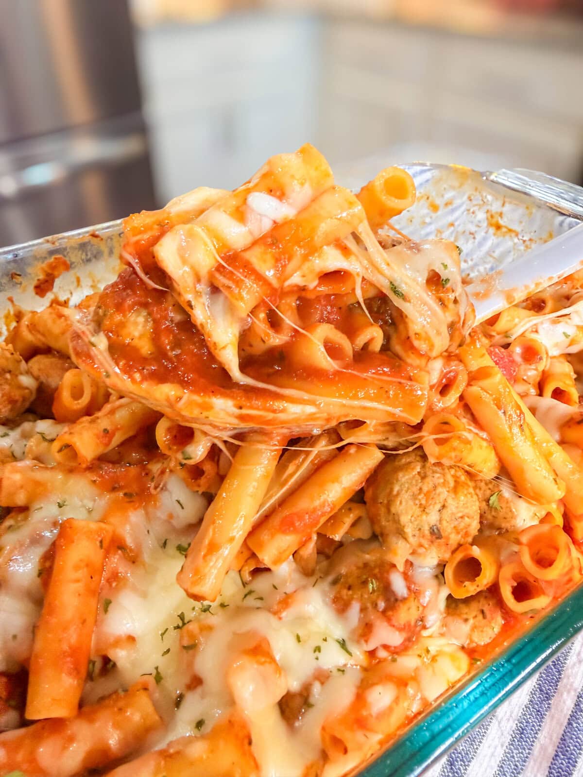 A close-up of One Dish Meatball Casserole with baked ziti, melted cheese, tomato sauce, and meatballs in a glass dish. A serving spoon lifts a portion, revealing gooey cheese and sauce. The background shows a blurred kitchen scene.