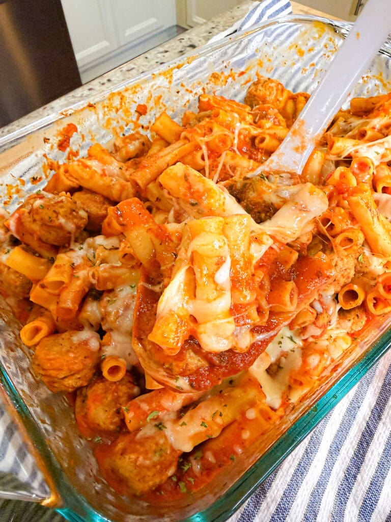A glass baking dish filled with One Dish Meatball Casserole—ziti pasta, meatballs, marinara sauce, and melted cheese—while a spatula lifts a portion. The dish rests on a striped cloth in a cozy kitchen setting.