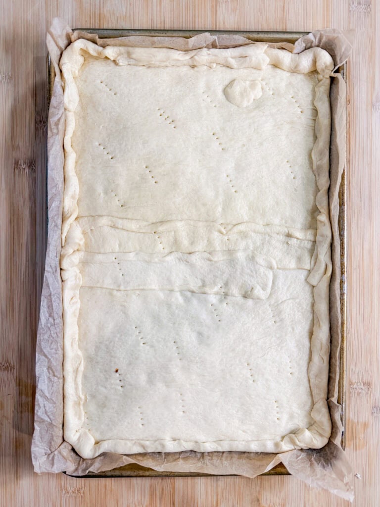 This Crescent Dough Veggie Pizza features a rectangular pastry with a lightly pricked, unbaked dough crust in a parchment-lined baking tray. The edges are neatly folded over along the borders, ready for topping.